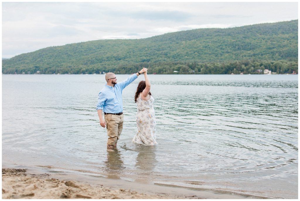 man twirling his fiance in the water overlooking Adirondack mountain range in Lake George, New York 
